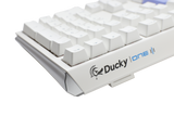 Close-up of a white Ducky One 3 mechanical keyboard with gray and white PBT keycaps on a light gray background. The Ducky logo is visible on the lower right side of the keyboard.