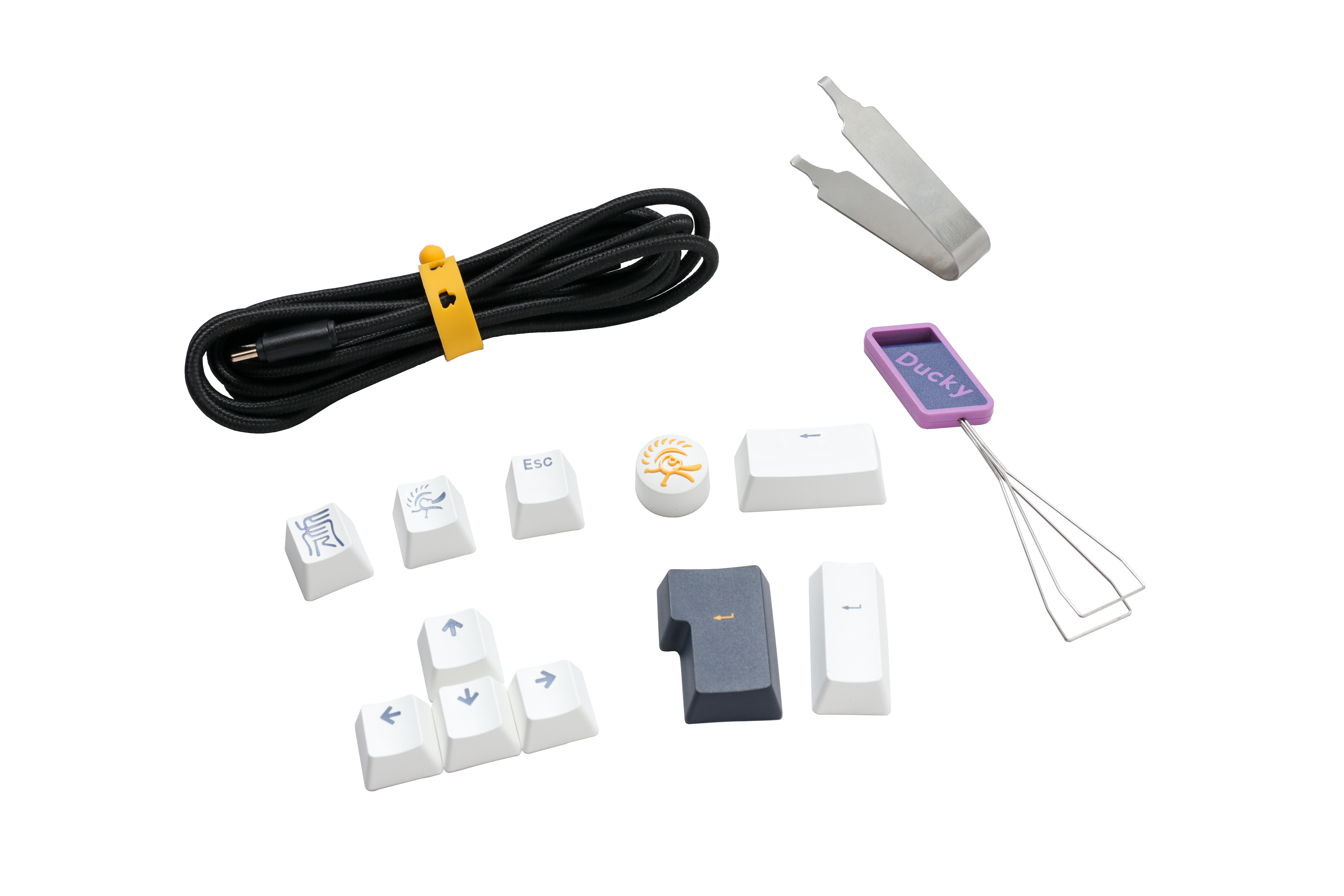 An assortment of Ducky keyboard accessories including a keycap puller, various PBT keycaps with symbols, a coiled cable, and additional small tools, all displayed on a white background.